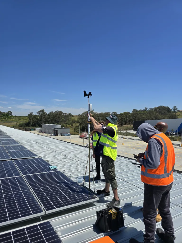 Two technicians on a commercial roof measuring airflow with a handheld anemometer beside solar panels