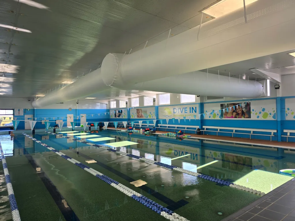 Indoor aquatic centre pool hall with a white fabric supply-air duct along the ceiling